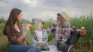 Happy family weekend, cheerful man feeding woman bun while relaxing on picnic with child drinking milk in green field