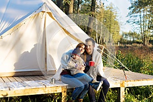 Happy family wraps blanket over themselves while sitting near canvas tent in the woods