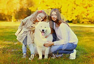 Happy family, young mother and child daughter with white Samoyed dog together in sunny autumn park