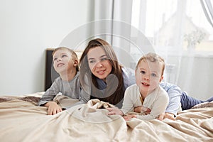 Happy family. Young mother dressed in light blue pajama lays with her two little sons on the bed with beige blanket in