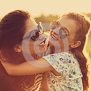 Happy fashion laughing kid girl embracing her mother with strong love in trendy sunglasses smiling and looking on nature