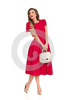 Happy Fashionable Woman In Red Dress, Gold High Heels And Beige Purse Is Looking Away