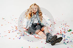 Happy fashionably dressed curly hair tween girl in in a denim jacket and black tutu skirt and rough boots sitting on white