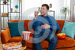 Happy fat man sitting on sofa eating junk food chips pizza popcorn and beer, watching TV film game