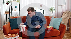 Happy fat man sitting on sofa eating junk food chips pizza popcorn and beer, watching TV film game