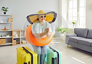 Happy fat woman in hat and sunglasses getting ready for holiday trip standing with suitcase at home