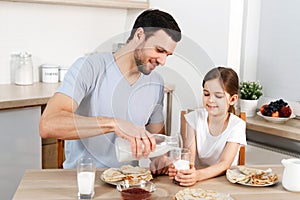 Happy father and daughter have breakfast at kitchen, eats delicious pancakes with jam, drink milk, enjoys delicious food