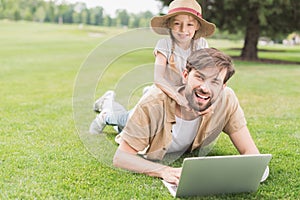 happy father and daughter using laptop and smiling at camera while lying on grass