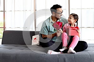 Happy Father Day, cute little girl sits on daddy lap on sofa in living room, giving present gift box flower and card to dad for