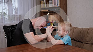 Happy father is engaged in arm wrestling with his son at home at the table.
