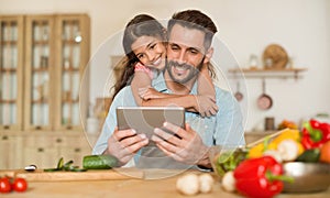 Happy father and his daughter using digital tablet, girl embracing dad from back while cooking together in kitchen