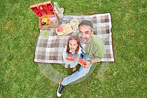 Happy father and his lovely little daughter smiling at camera, eating watermelon while having a picnic in the park