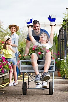 Happy father and his son playing with a wheelbarrow while girl and her mother looking them in the greenhouse.