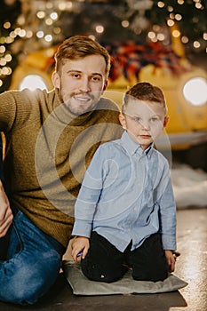 Happy father with his son sit on floor against background of glowing headlights and Christmas decorations