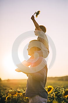 Happy father with his son on their shoulders having fun on a green field of blooming sunflowers at sunset. Hand up