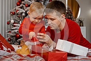 Happy Father and his sweet little son are very busy unwrapping red new year present lying on bed in front of them with Christmas