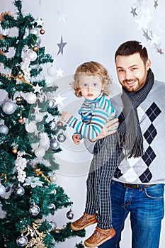 Happy father holds son near Christmas tree to decorate in white interior