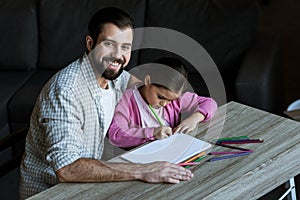 happy father with little daughter sitting at table and drawing in scrapbook