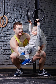 Happy father and little son exercising with gimnastic rings and smiling against brick wall in the gym.