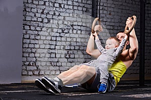 Happy father and little son exercising with gimnastic rings and smiling against brick wall in the gym.