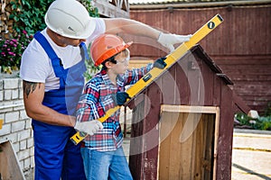 Happy father looking at son measuring wooden plank with building level. Building a dog house at backyard.