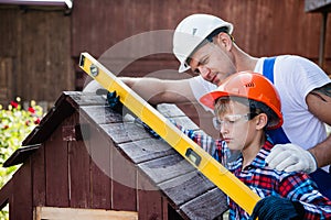 Happy father looking at son measuring wooden plank with building level. Building a dog house at backyard.