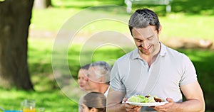 Happy father offering plate of food to camera at family barbecue