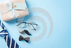 Happy Father`s Day background concept with blue necktie, glasses, black mustache and gift box on bright pastel background with