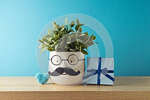 Happy Father's day concept with cute funny plant with mustache, lasses and gift box on wooden table over blue background
