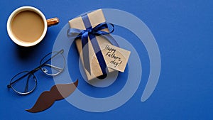 Happy Father`s day concept. Flat lay gift box with blue ribbon bow, male glasses, cup of coffee and mustache on blue background