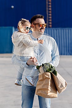 A happy father with a small daughter on his shoulders is walking in the parking lot holding paper bags in his hands with fresh veg