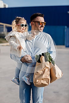 A happy father with a small daughter on his shoulders is walking in the parking lot holding paper bags in his hands with fresh veg