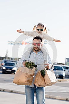 A happy father with a small daughter on his shoulders is walking in the parking lot holding paper bags in his hands with fresh veg