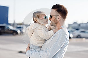 A happy father with a small daughter on his shoulders is walking in the parking lot holding paper bags in his hands with fresh veg