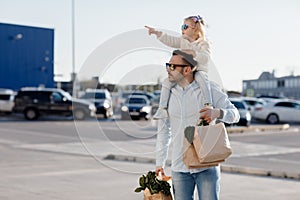 A happy father with a small daughter on his shoulders is walking in the parking lot holding paper bags in his hands with fresh veg