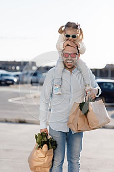 A happy father with a small daughter on his shoulders is walking in the parking lot holding paper bags in his hands with fresh veg