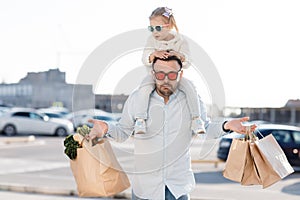 A happy father with a small daughter on his shoulders is walking in the parking lot holding paper bags in his hands with fresh veg