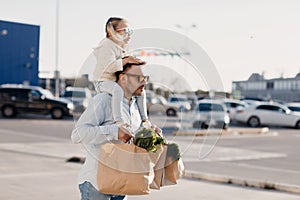 A happy father with a small daughter on his shoulders is walking in the parking lot holding paper bags in his hands with fresh veg