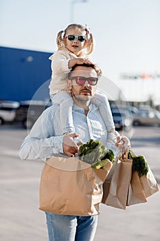 A happy father with a small daughter on his shoulders is walking in the parking lot holding paper bags in his hands with fresh veg