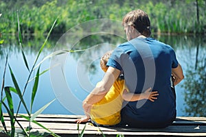 Happy father and son hugging sitting on wooden pier near lake on sunny summer day. Dad and child boy spend time together on wharf
