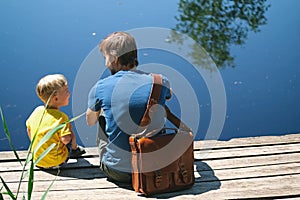 Happy father and son sitting on wooden pier near lake on summy summer day. Dad and child boy spend time together on wharf