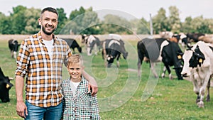 happy father and son smiling at camera while standing near grazing cattle