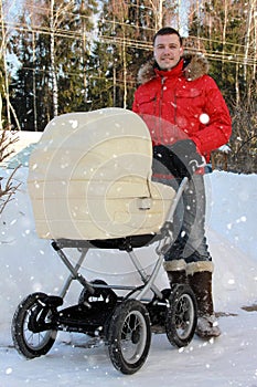 happy father stands with pram with sleeping child in backyard with snowdrifts on winter sunny day. Man in red jacket looks into