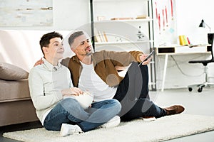 Happy father and teen son watching tv and holding bowl of popcorn on floor