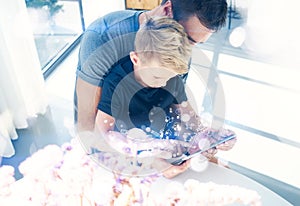 Happy father with young son using tablet PC in sunny room.Dad and little boy playing mobile computer, resting indoor