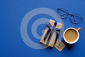 Happy Fathers day concept. Top view gift box wrapped blue ribbon, greeting card, cup of coffee and glasses on blue desk. Father`s
