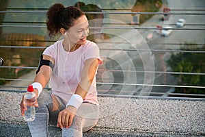 Happy female athlete sportswoman runner jogger in sportswear holding a water bottle and resting on city bridge after morning