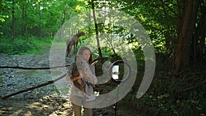 Happy female blogger recording video through mobile phone and ring lamp with dog and horses on background outdoor.