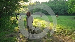 Happy female blogger recording video through mobile phone and ring lamp with dog and horses on background outdoor.