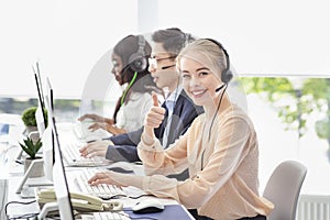 Happy female call centre consultant showing thumb up gesture and her colleages at modern office